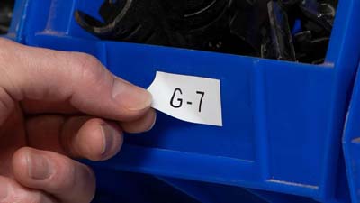 A white, repositionable vinyl label with the text G-7 printed on it and applied to a bin in a warehouse. A worker is peeling it off to move it from one bin to another.
